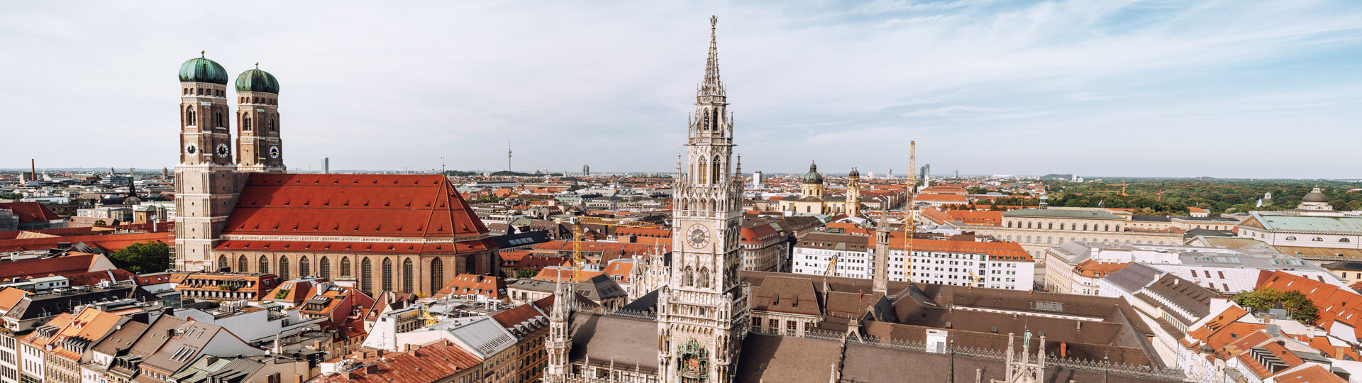 Der Marienplatz in München ist von historischen Gebäuden umgeben; im Vordergrund steht dasNeue Rathaus mit seinem charakteristischen Glockenturm, während im Hintergrund die Türme der Frauenkirche sichtbar sind. Der Platz ist belebt, und der Himmel ist blau mit wenigen Wolken.